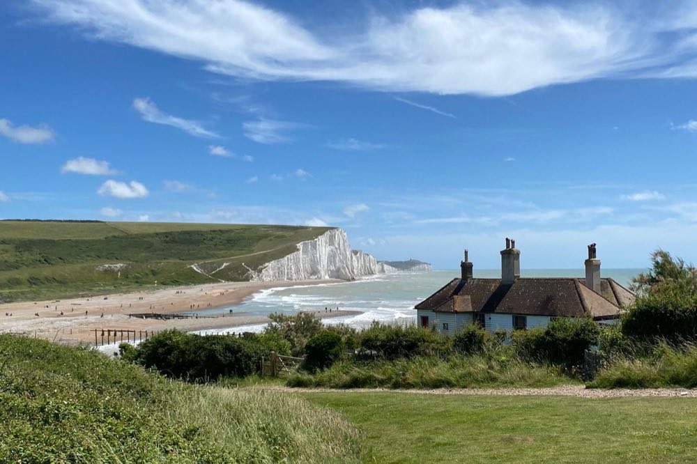 The white chalk cliffs of the Seven Sisters beyond a shingle beach and a clifftop cottage, on a sunny day